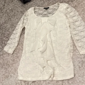 Vit spetsblus med rosett och volang - Säljer en vit spetsblus med trekvartsärm. Blusen har en stor dekorativ rosett och volang framtill, samt rundad halsringning. Perfekt för dig som gillar romantiska detaljer.