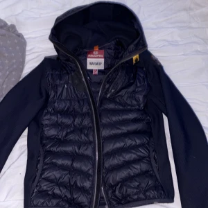 Svart hybridjacka från Parajumpers - Säljer en svart hybridjacka från Parajumpers med quiltade partier framtill och baktill samt huva. Jackan har dragkedja och broderad logotyp på ärmen. Perfekt för kyliga dagar.