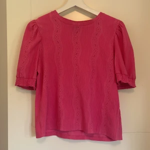 Rosa blus med puffärm - Säljer en söt rosa blus med korta puffärmar och vågigt hålmönster. Blusen har rynkade ärmslut och en rund halsringning. Perfekt för dig som vill ha en färgglad och feminin topp.