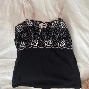 Gullig vintage topp!! - Säljer en svart linnetopp med tunna axelband och vacker spets upptill. Toppen har broderade blommor i vitt och ljusrosa samt dekorativ snörning framtill🌸💞