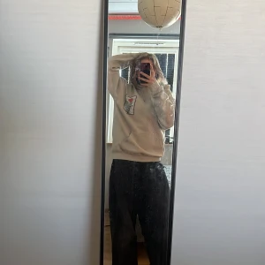 Beige hoodie med japanskt tempeltryck - Säljer en beige hoodie med stort tryck på ryggen föreställande ett japanskt tempel, röd sol och blå himmel. På bröstet finns en mindre version av samma motiv. Hoodien har huva och långa ärmar. Perfekt för dig som gillar streetwear och unika prints. 