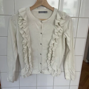 Vit kofta med volang från Åhléns - Säljer en söt vit kofta från Åhléns med långa ärmar och knappar framtill. Koftan har dekorativa volanger längs båda sidor av knäppningen och är stickad i mjukt material. Perfekt för dig som gillar romantiska detaljer!