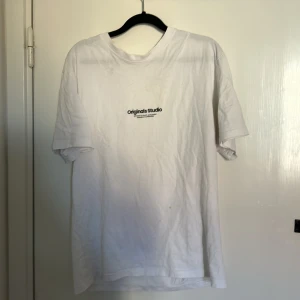 Vit t-shirt från Originals Studio - Säljer en klassisk vit t-shirt från Originals Studio med liten svart logga på bröstet. T-shirten har rund hals och korta ärmar, perfekt för en clean och enkel stil.