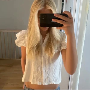 Vit Zara blus - Säljer en söt vit blus med korta volangärmar och knappar framtill. Blusen är croppad och har fina veckade detaljer på framsidan. Perfekt för en fräsch och somrig look. Storleken är 10år men jag hade den när jag va 12år så skulle säga att man kan ha den ändå! 🥰