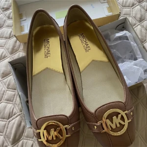 Bruna ballerinaskor från Michael Kors med gulddetaljer - Säljer ett par snygga bruna ballerinaskor från Michael Kors med rund tå och stora guldfärgade MK-loggor framtill. Skorna har platt sula och är perfekta till vardagen eller fest. Köpte dessa men tyvärr så passade de inte mig. De ska vara storlek 39 men var för små på mig.  Ny Pris: 1300 kr.  Finns i Västerås.  