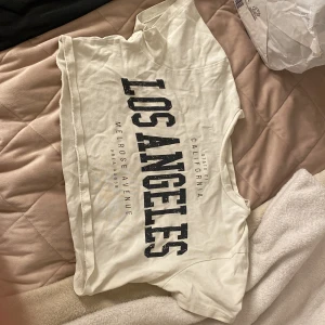 Vit croppad t-shirt med Los Angeles-tryck - Säljer en vit croppad t-shirt med stort svart tryck 'Los Angeles' på framsidan. T-shirten har korta ärmar och rund halsringning. Perfekt för en varm sommarkväll och man känner att man inte vill bli för varm☀️