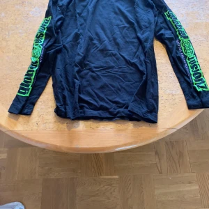 Svart långärmad tröja från H&M med neontryck - Cool svart långärmad tröja från H&M med grönt och blått tryck på båda ärmarna där det står 'global vision' och en jordglob. Tröjan är i mjuk bomull och har en avslappnad passform. Perfekt för dig som gillar streetwear och tydliga detaljer.