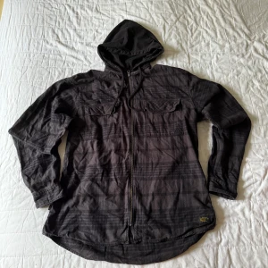 Svart overshirt med huva och dragkedja - Säljer en svart overshirt med huva och dragkedja framtill. Skjortan har två bröstfickor med lock och ett diskret rutigt mönster. Perfekt för lager-på-lager och streetwear-stil.