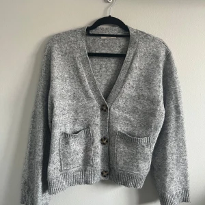 Grå stickad kofta från Gina Tricot - Mysig grå stickad kofta från Gina Tricot med två stora fickor framtill och knappar i sköldpaddsmönster. Koftan har v-ringning och ribbade muddar vid ärmslut och nederkant. Perfekt för lager-på-lager under kyliga dagar. Pris kan diskuteras🩷 tvättar såklart innan jag skickar!🩷 använd några fåtal gånger. Har inga hål eller fläckar!🩷