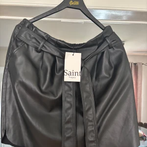 Svart kjol i skinnimitation från Saint Tropez - Säljer en svart kjol i skinnimitation från Saint Tropez. Kjolen har ett bälte i midjan och breda hällor. Snygg och stilren design som passar perfekt till många olika looks.