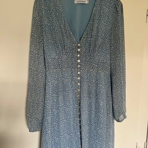 Blå mönstrad långklänning från Adoore - Säljer en blå klänning från Adoore med vitt mönster och v-ringning i modellen Paris. Klänningen har långa ärmar och knappar framtill hela vägen ner. Perfekt för vår och sommar, med en feminin och tidlös look. knappen längst ner saknas, men inget som påverkar funktionen. 