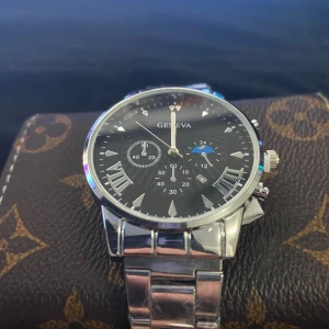 Silverfärgad klocka  - Stilren klocka från  med svart urtavla, romerska siffror och detaljer i silver. Klockan har en länk i metall och flera små urtavlor för extra funktioner. Perfekt accessoar för dig som gillar klassisk design.