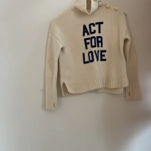 Vit polotröja med text och knappar - Säljer en vit polotröja med blå texten 'ACT FOR LOVE' framtill. Tröjan har ribbade muddar, hög krage och dekorativa knappar på axeln. Perfekt för dig som gillar statement-plagg och vill ha något unikt i garderoben. Använd jätte få gånger