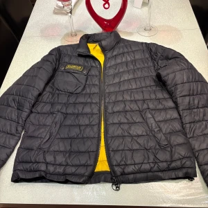 Svart quiltad pufferjacka från Barbour - Säljer en svart quiltad pufferjacka från Barbour med gul insida och dragkedja. Jackan har en bröstficka med Barbour-logga och två sidofickor. Perfekt för kyliga dagar och har en modern look.