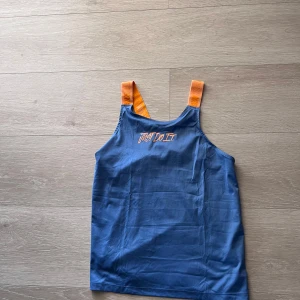 Blå linnetopp med orange axelband - Säljer en blå linnetopp med orange axelband och texten 'Just Do It' broderad framtill. Toppen har en sportig känsla och är perfekt för varma dagar eller träning.