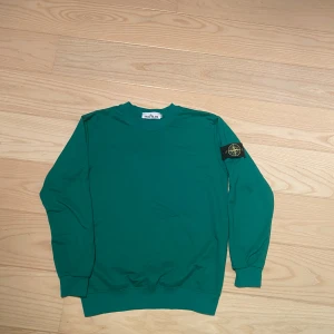 Grön sweatshirt från Stone Island - Säljer en grön sweatshirt från Stone Island med klassisk patch på vänster ärm. Tröjan har rund halsringning och ribbade muddar vid ärmslut och nederkant. Perfekt för en stilren och avslappnad look. Den är i riktigt bra skick. Storlek M men sitter lite mindre. Skriv om ni har några funderingar!