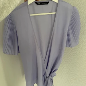 Ljuslila omlottblus från Zara - Säljer en ljuslila omlottblus från Zara med korta, plisserade ärmar och knyt i midjan, vilket gör den justerbar i midjan och gör att den passar flera storlekar💕Perfekt för vår och sommar!