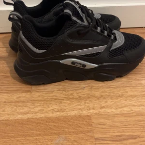 Svarta sneakers från Dior med grå detaljer - Säljer ett par svarta sneakers från Dior med grå och silverfärgade detaljer. Skorna har snörning, meshpartier och en chunky sula. Perfekta för dig som gillar en sportig och modern stil.