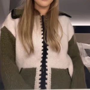 Stickad Zara cardigan i vitt och olivgrönt - Säljer en mysig stickad cardigan jacka med vita och olivgröna partier samt svarta detaljer längs dragkedjan och kragen. Jackan har långärmade ärmar och en avslappnad passform. Perfekt för lager-på-lager-stil.