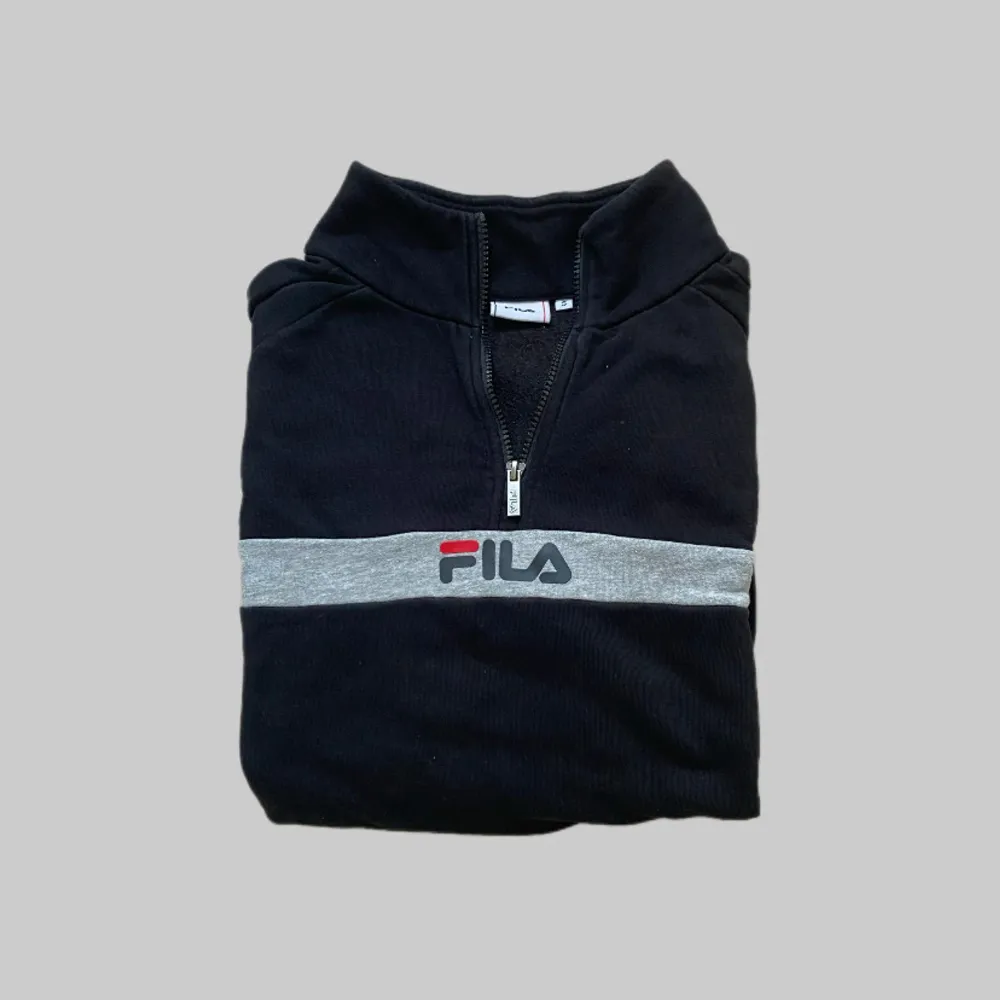 Svart tröja från FILA med halv dragkedja och grått band över bröstet med broderad logga. Perfekt för en sportig och avslappnad stil. Mjuk bomullskänsla och hög krage.. Neuletakit & Villapaidat.