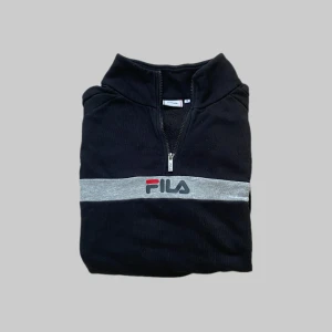 Svart ziptröja från FILA - Svart tröja från FILA med halv dragkedja och grått band över bröstet med broderad logga. Perfekt för en sportig och avslappnad stil. Mjuk bomullskänsla och hög krage.