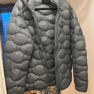 Grå dunjacka från Peak Performance - Säljer en grå dunjacka från Peak Performance med huva och dragkedja. Jackan har ett vågformat quiltat mönster och två sidofickor. Perfekt för kyliga dagar och enkel att matcha med olika outfits. Har används 1 gång. Pris kan gå ner med snabb affär