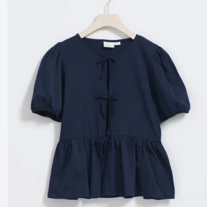 Mörkblå blus med knyt - Supersöt mörkblå blus med korta puffärmar och knyt framtill. Blusen har en lös passform och volang nedtill som ger en avslappnad look. Perfekt att styla med jeans eller kjol!