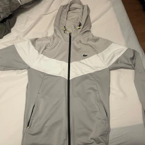 Grå och vit kofta från Lacoste Sport - Snygg grå och vit kofta från Lacoste Sport med huva och dragkedja. Jackan har en sportig design med vit panel över bröstet och klassisk Lacoste-logga på vänster sida. Justerbar huva med snörning och två fickor med dragkedja.