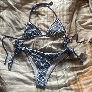Blåvit bikini med mönster och knyt - Säljer en snygg bikini i storlek S med blått och vitt blommigt mönster. Både överdel och underdel har knytband i sidorna och nacken. Materialet är mjukt och stretchigt, tillverkat av polyester och elastan. Perfekt för stranden eller poolen!