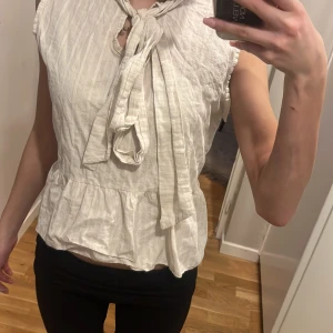 Vit blus med knytband - Säljer en söt vit blus med knytband i halsen och volang nertill. Blusen är ärmlös och har ett diskret mönster i tyget. Perfekt för dig som gillar feminina detaljer och en luftig känsla.efter som det är fläckar kan priset dikteras stryks innan jag skickar