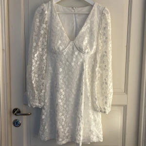Studentklänning vit blommig kortklänning  - Superfin vit kortklänning med v-ringning och långa ärmar. Klänningen har ett vackert blommigt spetsmönster och markerad byst. Perfekt för student eller sommar. 