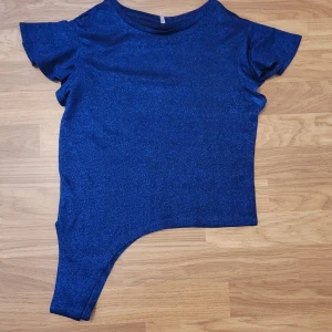Blå glittrig tröja - Säljer en blå glittrig tröja med korta ärmar och rund halsringning. Tyget har en skimrande finish som gör den perfekt för att sticka ut. Asymmetrisk nederdel som ger en unik look.