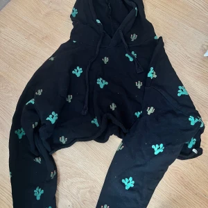 Svart hoodie med kaktusmönster från Pull&Bear - Säljer en svart hoodie från Pull&Bear med gröna och beige kaktusar broderade över hela tröjan. Tröjan har huva med dragsko och långa ärmar. Perfekt för dig som gillar unika detaljer och skön stil.