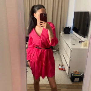 Rosa v-ringad klänning med volang - Säljer en knallrosa klänning med v-ringning och långa ärmar. Klänningen har volangdetalj nertill och en lös passform, men kan stylas med bälte i midjan. Perfekt för sommarens tillfällen!