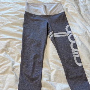Gråa leggings med hög midja och vit logga - Säljer ett par gråa leggings med bred, ljusgrå hög midja och stor vit logga längs benet. Perfekta för träning eller chill. Mjukt och stretchigt material som sitter skönt hela dagen.