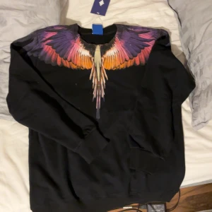 Marcelo Burlon - Cool svart sweatshirt från Marcelo Burlon med ett stort färgglatt vingmönster i lila, rosa, orange och gult över axlar och bröst. Tröjan har långa ärmar och klassisk passform. Perfekt statement-plagg för dig som vill sticka ut. Helt ny