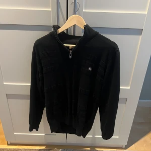 Svart stickad ziptröja från Burberry - Säljer en svart stickad ziptröja från Burberry med diskret logga på bröstet. Tröjan har hög krage, dragkedja framtill och ribbade muddar. Perfekt för dig som gillar klassisk stil med exklusiv känsla.