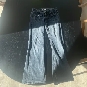 Blå bootcut jeans med fickdetaljer - Säljer ett par blå bootcut jeans med snygga fickor framtill med lock och knapp. Jeansen har klassisk femficksdesign och är i ett mjukt denimtyg. Perfekta för dig som gillar en lite vidare passform nertill.