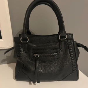 Svart handväska med dragkedjedetaljer - Snygg svart handväska i skinnimitation med dubbla handtag och dekorativa sömmar på sidorna. Väskan har flera fack med dragkedja både fram och bak samt inuti, perfekt för att organisera dina saker. Stilren och passar till många outfits. Pris går att diskutera!