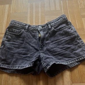 Svarta jeansshorts med hög midja - Säljer ett par svarta jeansshorts med hög midja och klassisk femficksdesign. Shortsen har dragkedja och knapp framtill samt synliga sömmar. Perfekta för varma dagar och enkel att matcha med olika toppar.