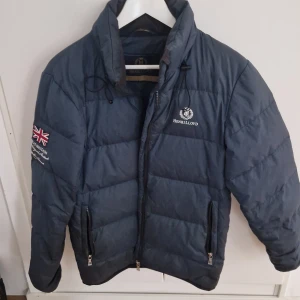 Blå pufferjacka från Henri Lloyd dunjacka - Säljer en blå pufferjacka från Henri Lloyd med broderad logga på bröstet och brittisk flagga på ärmen. Jackan har dragkedja framtill och två fickor med dragkedja. Perfekt för kyligare dagar.