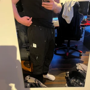 Svarta Adidas träningsbyxor - Säljer ett par svarta träningsbyxor som är som överdragsbyxor fast tunna från Adidas Originals med elastisk midja och dragsko. Byxorna har flera fickor med dragkedja och Adidas-logga på ena benet. Perfekta för en avslappnad streetwear-look. Knappast använda och i toppskick!