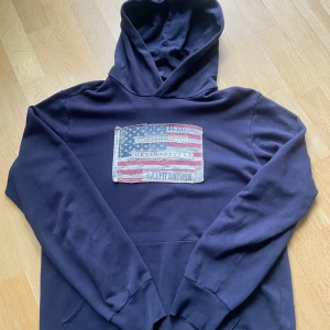 Mörkblå hoodie från Ralph Lauren - Säljer en mörkblå hoodie från Ralph Lauren Denim & Supply med en stor amerikansk flagga-tryck på bröstet. Tröjan har huva och en klassisk känguruficka framtill. Perfekt för en avslappnad stil.