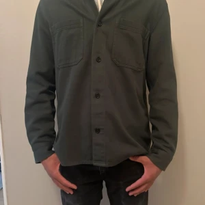 Mörkgrön overshirt med bröstfickor - Säljer en mörkgrön overshirt med två bröstfickor och knäppning framtill. Skjortan har lång ärm och en rak passform, perfekt för en stilren och enkel look. Passar dig som gillar klassiska plagg med diskreta detaljer.