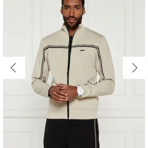 Beige track jacket från BOSS med svarta detaljer - Snygg beige track jacket från BOSS med svarta linjer längs ärmar och bröst. Jackan har dragkedja hela vägen, hög krage och diskret BOSS-logga på bröstet. Perfekt för en sportig och stilren look.