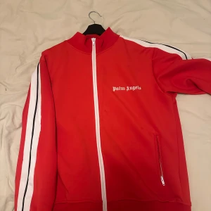 Röd track jacket från Palm Angels - Palm Angels röd track jacket med vita och svarta ränder längs ärmarna. Jackan har dragkedja framtill, ståkrage och två fickor med dragkedja. Ikonisk logga på bröstet. Perfekt för en sportig och trendig look.