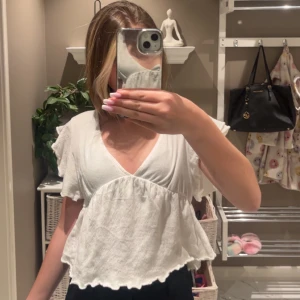 Vit v-ringad blus med volang - Säljer en söt vit blus med v-ringning och volangdetaljer. Toppen har korta ärmar och en lös passform med vågig nederkant. Den har blivit för liten för mig därav säljer jag den!❣️