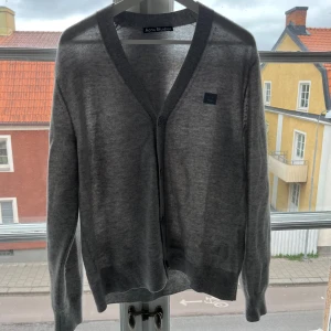 Grå kofta från Acne Studios - Säljer en stilren grå kofta från Acne Studios med v-ringning och knäppning framtill. Koftan har långa ärmar och en liten svart logotyp på bröstet. Perfekt för lager-på-lager och enkel att matcha.