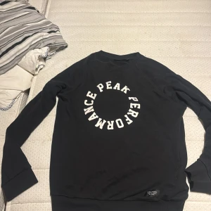 Svart sweatshirt från Peak Performance - Säljer en svart sweatshirt från Peak Performance med stort vitt tryck i cirkelformad text på bröstet. Tröjan har rund halsringning och långa ärmar. Perfekt för en avslappnad och sportig stil.
