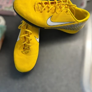 Gula Nike fotbollsskor med snörning - Säljer ett par gula Nike fotbollsskor med vit swoosh och svarta detaljer. Skorna har snörning och en platt sula med mönster för bra grepp. Perfekta för inomhusspel och har en sportig look.